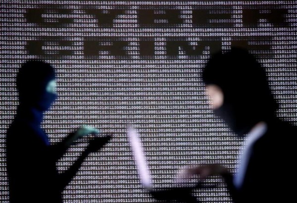 People wearing balaclavas are silhouetted as they pose with a laptops in front of a screen projected with the word cyber crime.