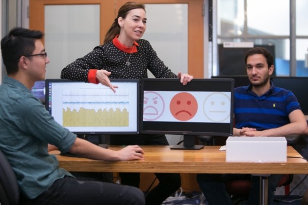 Professor Dina Katabi (center) explains how PhD student Fadel Adib's (right) face is neutral but that EQ-Radio's analysis of his heartbeat and breathing show that he is sad.